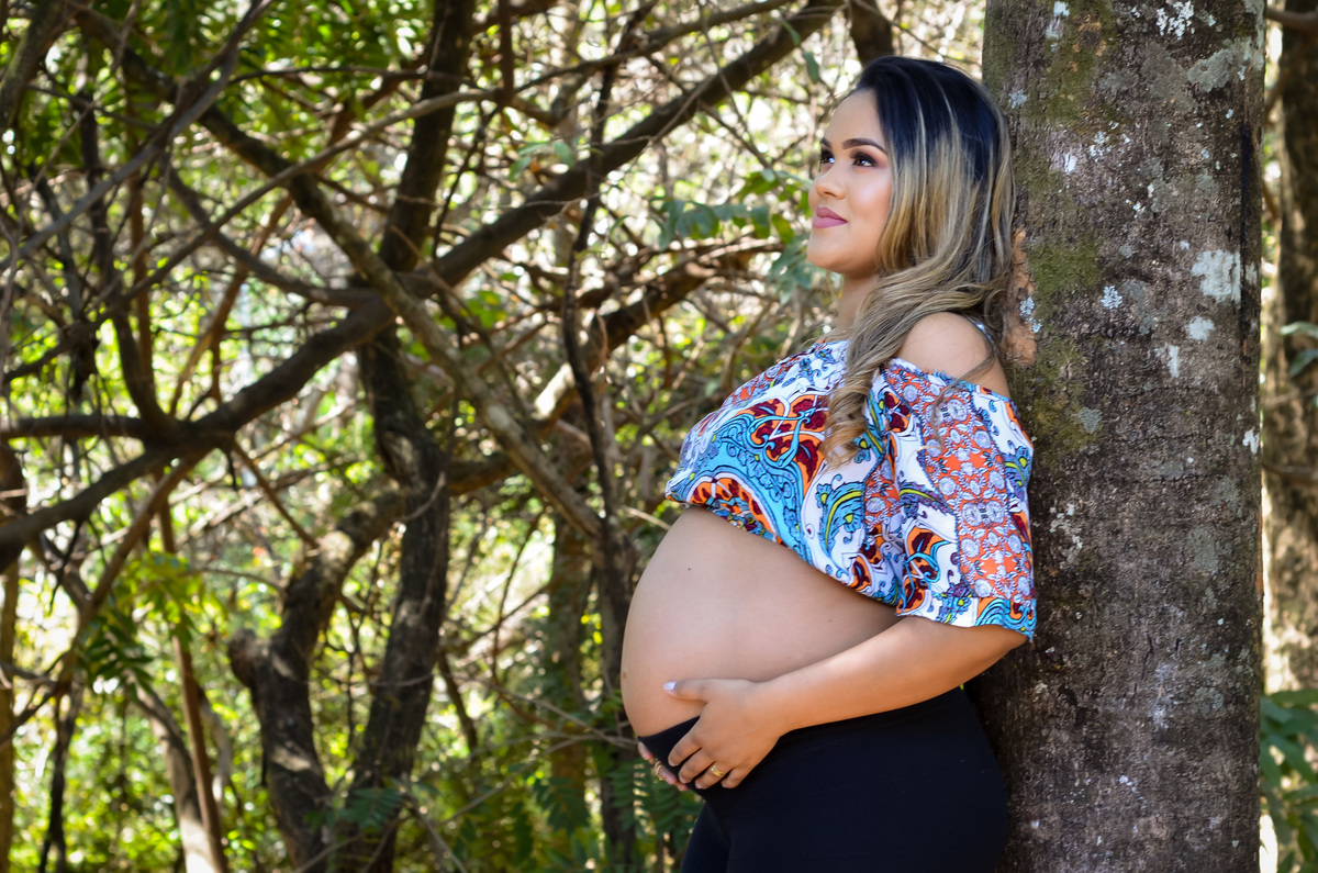 Foto gestante segurando a barriga encostada em uma árvore em meio ao verde 