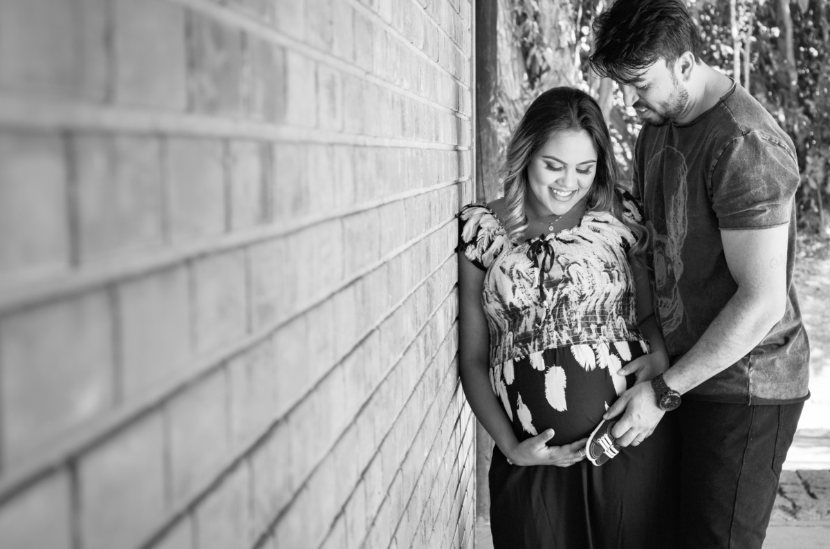 Foto casal gestante abraçados sorrindo encostados em uma parede de tijolinhos em um dia ensolarado em meio a árvores