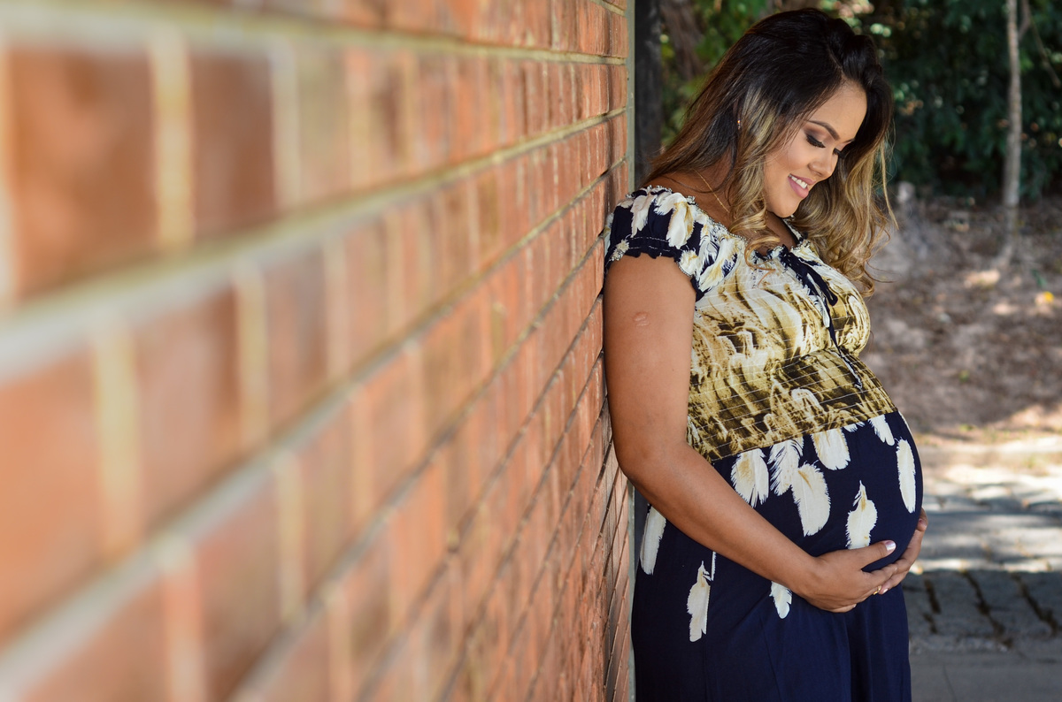 Foto gestante segurando e olhando para a barriga sorrindo encostados em uma parede de tijolinhos em um dia ensolarado em meio a árvores