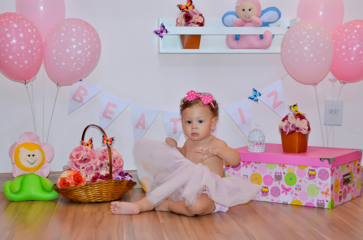 Foto de menina sentada com tutu rosa em cenário com flores e bichinhos de pelúcia 