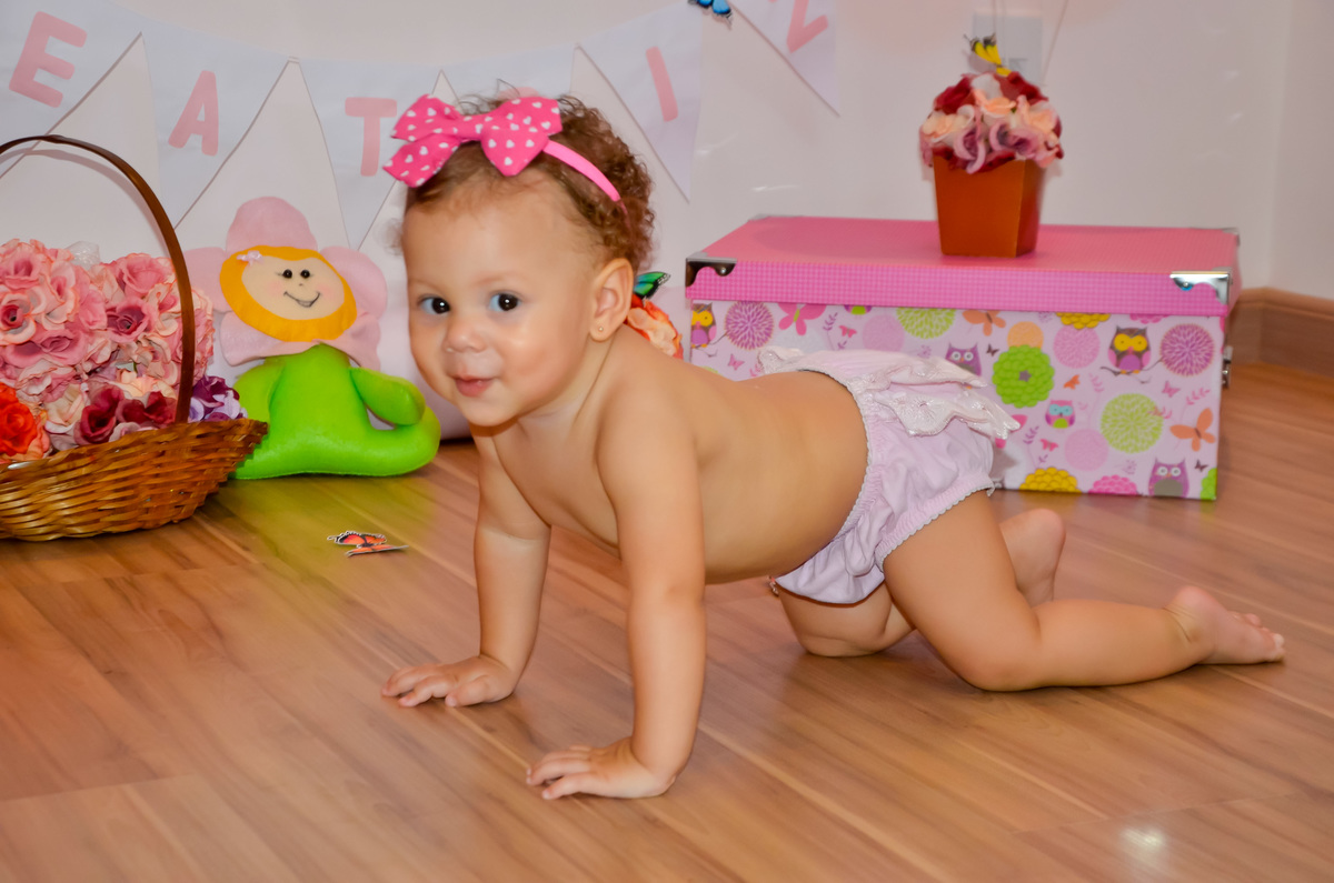 Foto de menina engatinhando e sorrindo em cenário com flores e bichinhos de pelúcia 