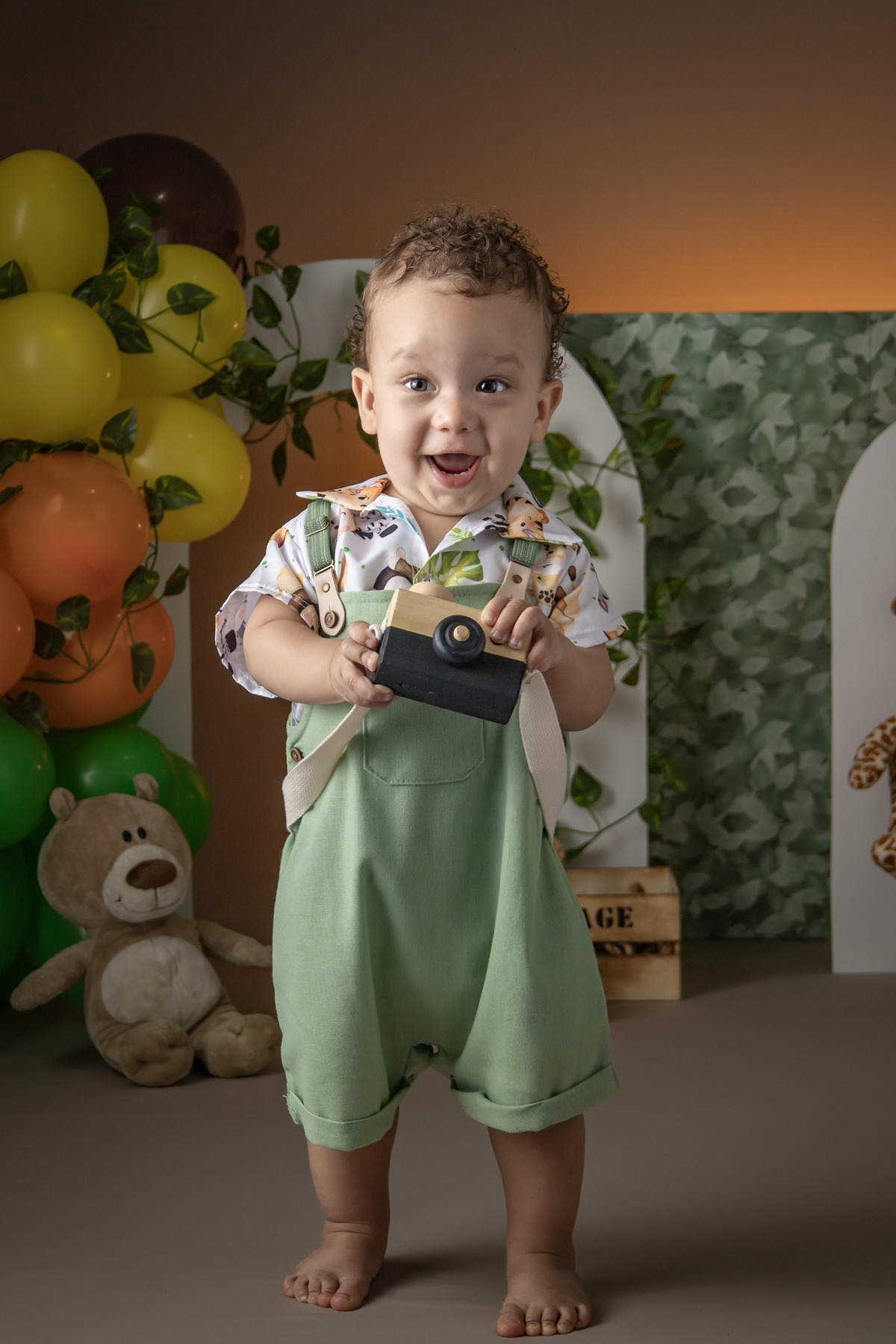 Ensaio fotografico infantil menino sorrindo, vestindo jardineira verde, segurando uma camera fotografica de brinquedo, tema safari 