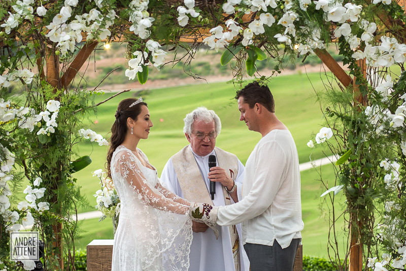 Os noivos dão as mãos enquanto o Padre pede a bênção de Deus sobre as alianças, em fotografia de Andre Attene