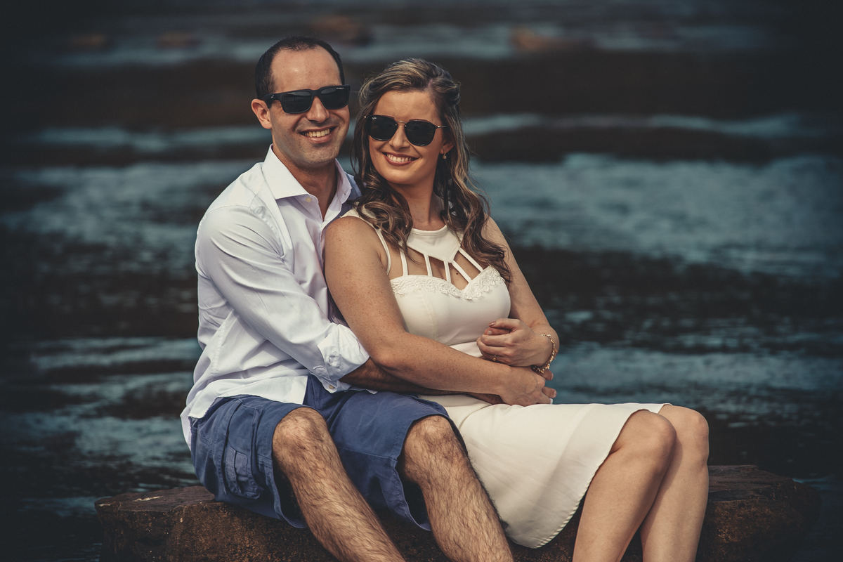 Fotógrafo de casamento de Cachoeirinha fotografia ensaio pré casamento casal abraçado sentado na pedra na beira do rio com óculos escuros