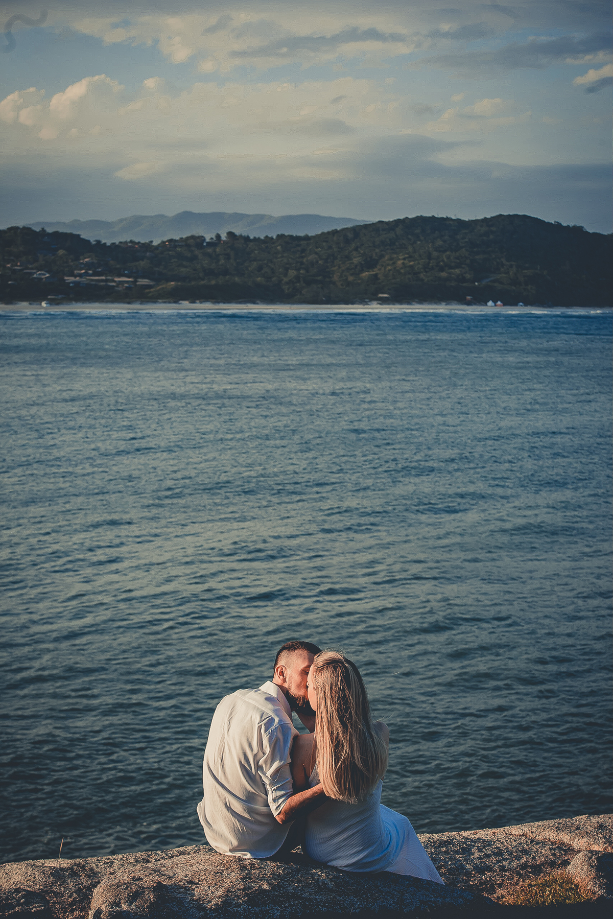Fotógrafo de casamento de Cachoeirinha fotografia ensaio pré casamento casal de costas se beijando sentado nas pedras com o mar ao fundo