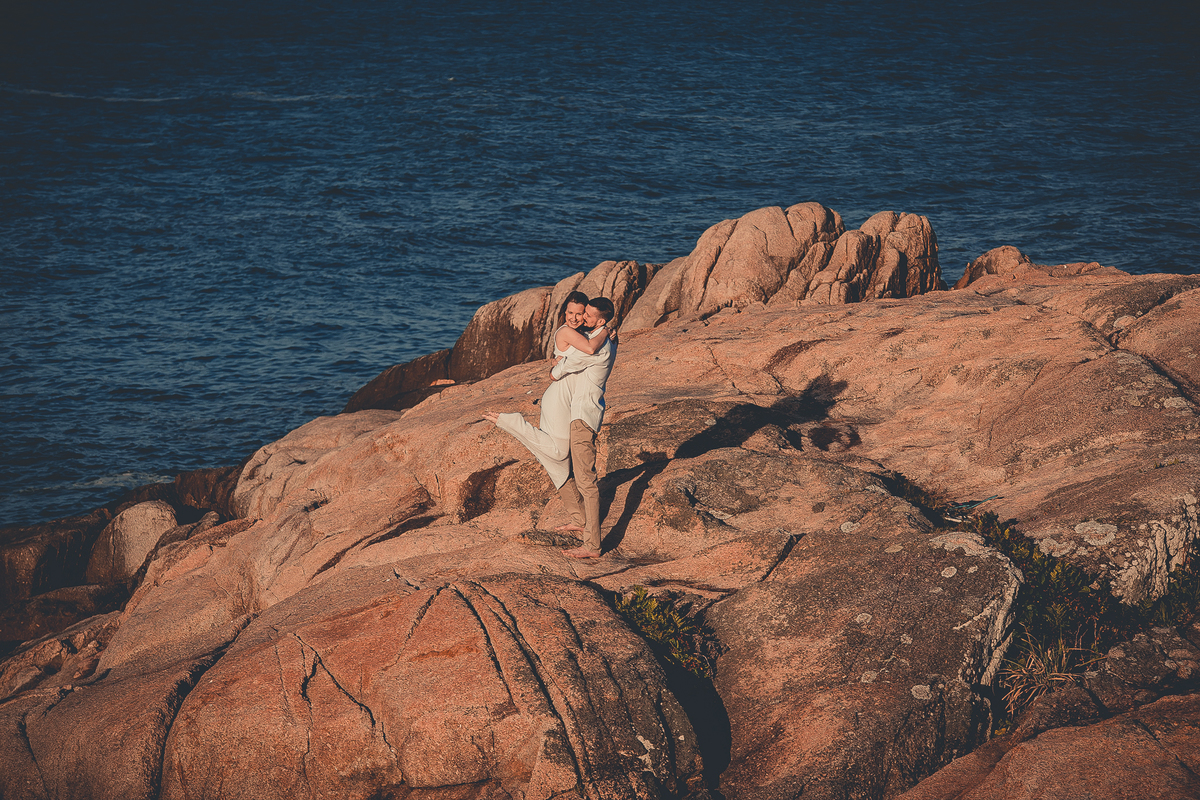 Fotógrafo de casamento de Cachoeirinha fotografia ensaio pré casamento casal nas pedras e o mar ao fundo ela no colo dele