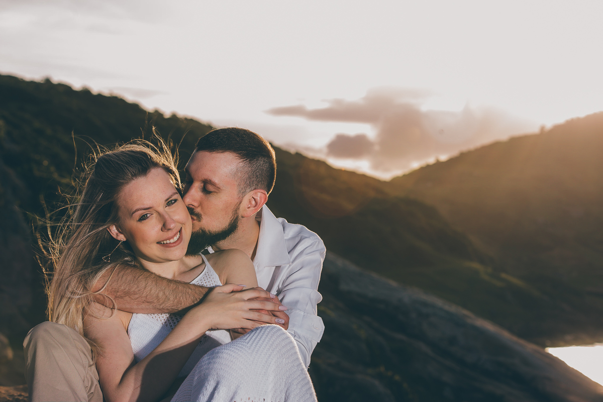 Fotógrafo de casamento de Cachoeirinha fotografia ensaio pré casamento casal sentado nas pedras no morro ele beijando ela e ela sorrindo imagem fechada