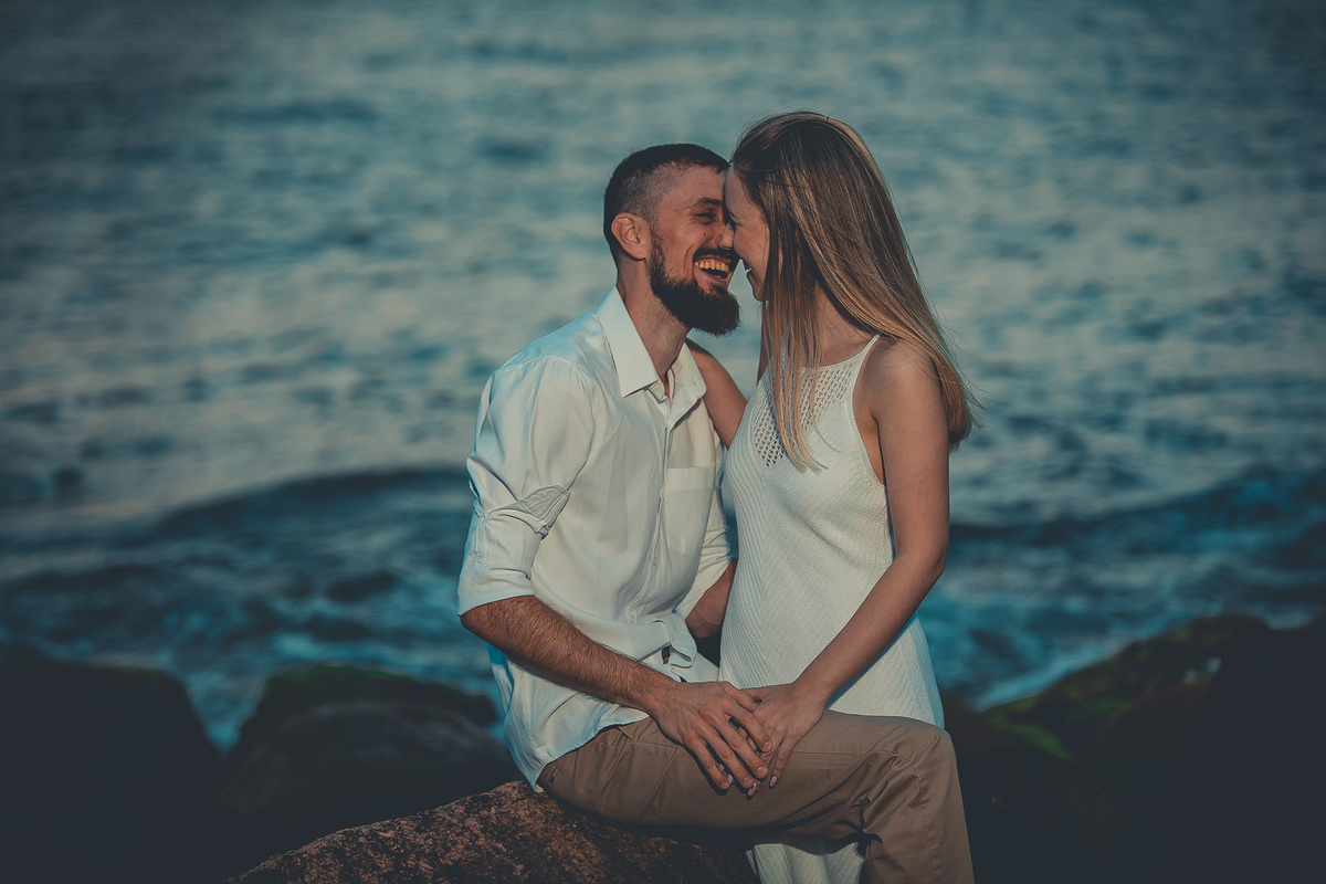 Fotógrafo de casamento de Cachoeirinha fotografia ensaio pré casamento casal sorrindo um para o outro sentado nas pedras e o mar ao fundo