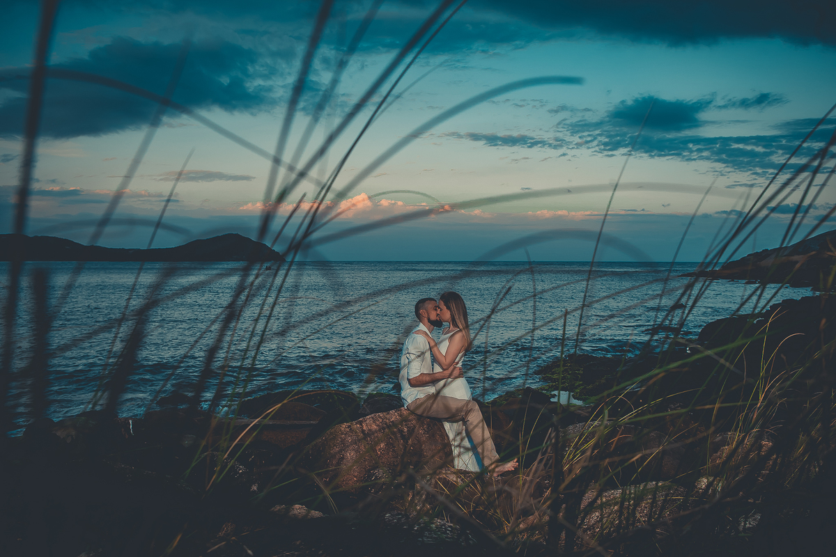 Fotógrafo de casamento de Cachoeirinha fotografia ensaio pré casamento casal se beijando sentado nas pedras o mar ao fundo e imagem entre os arbustos