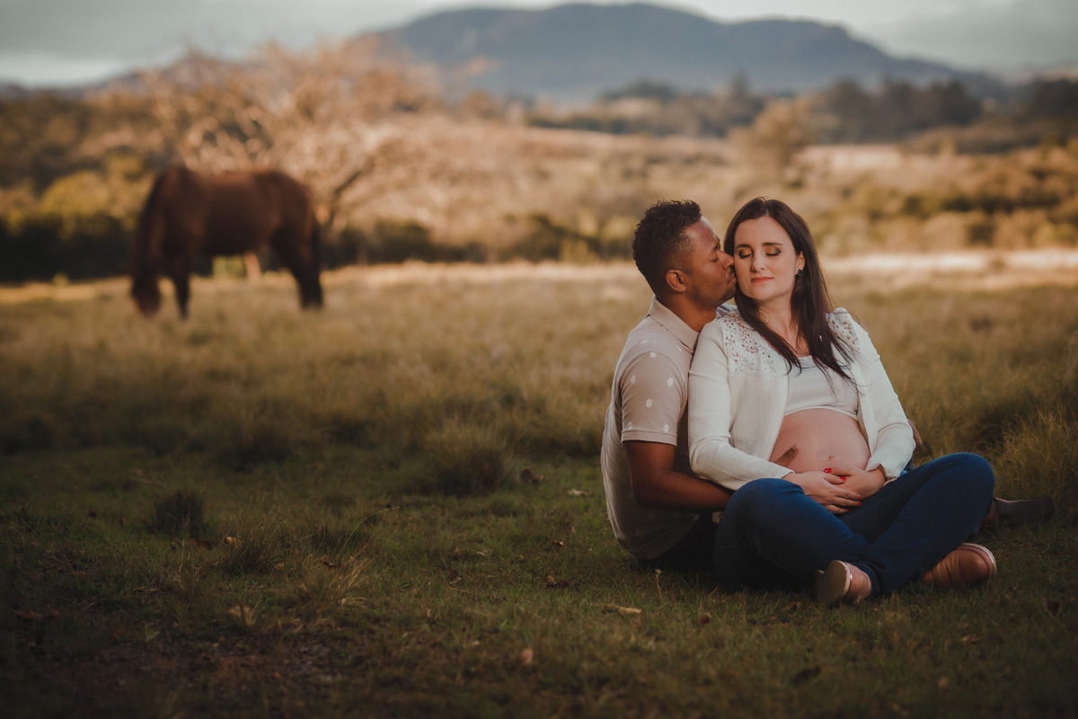 book de gestante com casal sentado um do lado do outro e esposo dando beijo na esposa e com cavalo ao fundo