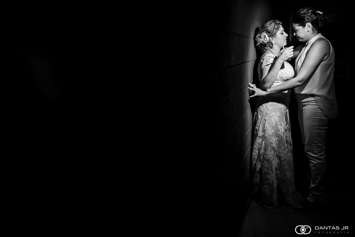 Casal de mulheres lésbicas em casamento homoafetivo se beijando encostadas em parede sob um feixo de luz por Dantas Jr. Fotografia