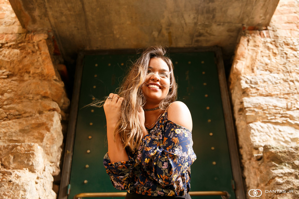 Ensaio debutante com menina olhando a paisagem e sorrindo enquanto cabelos voam em frente a porta de metal no Parque das Ruínas em Santa Tereza RJ por Dantas Jr. Fotografia