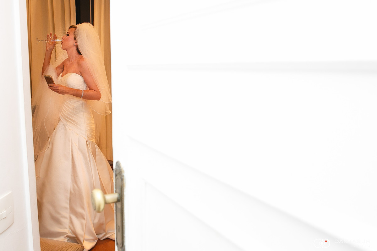 Noiva observada em fresta na porta durante making of enquanto bebe uma taça de champagne na casa do alto por Dantas Jr.
