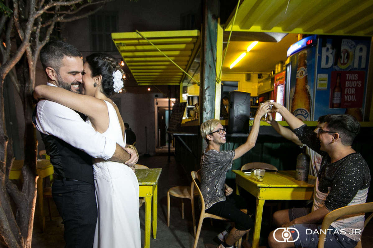 Casal de noivos se beijando em frente a boteco enquanto fregueses brindam clicados por Dantas Jr