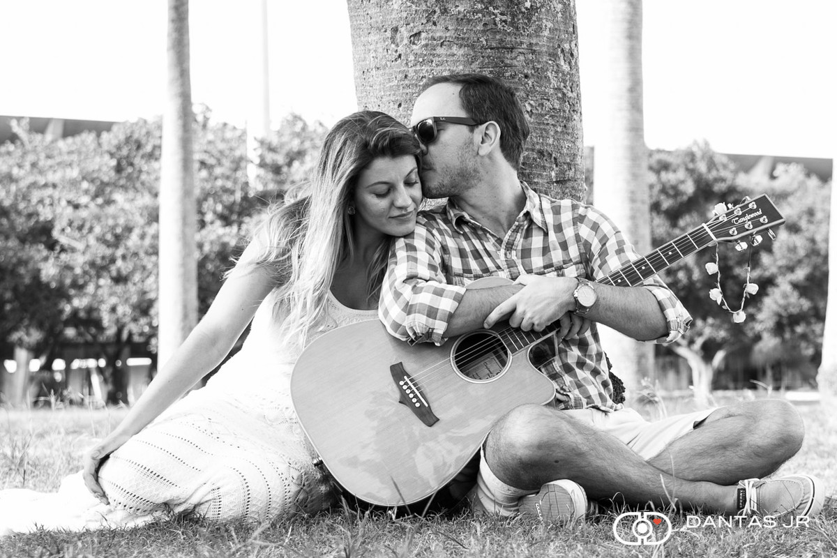 Casal namorando enquanto ele toca violão de oculos escuros e dá um beijo na cabeça dela