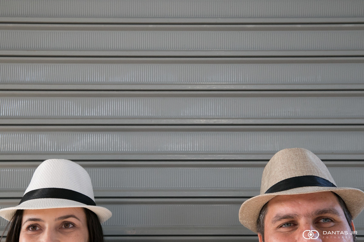 Casal mostrando apenas os olhos e com chapéu panamá encostados numa porta de alumínio