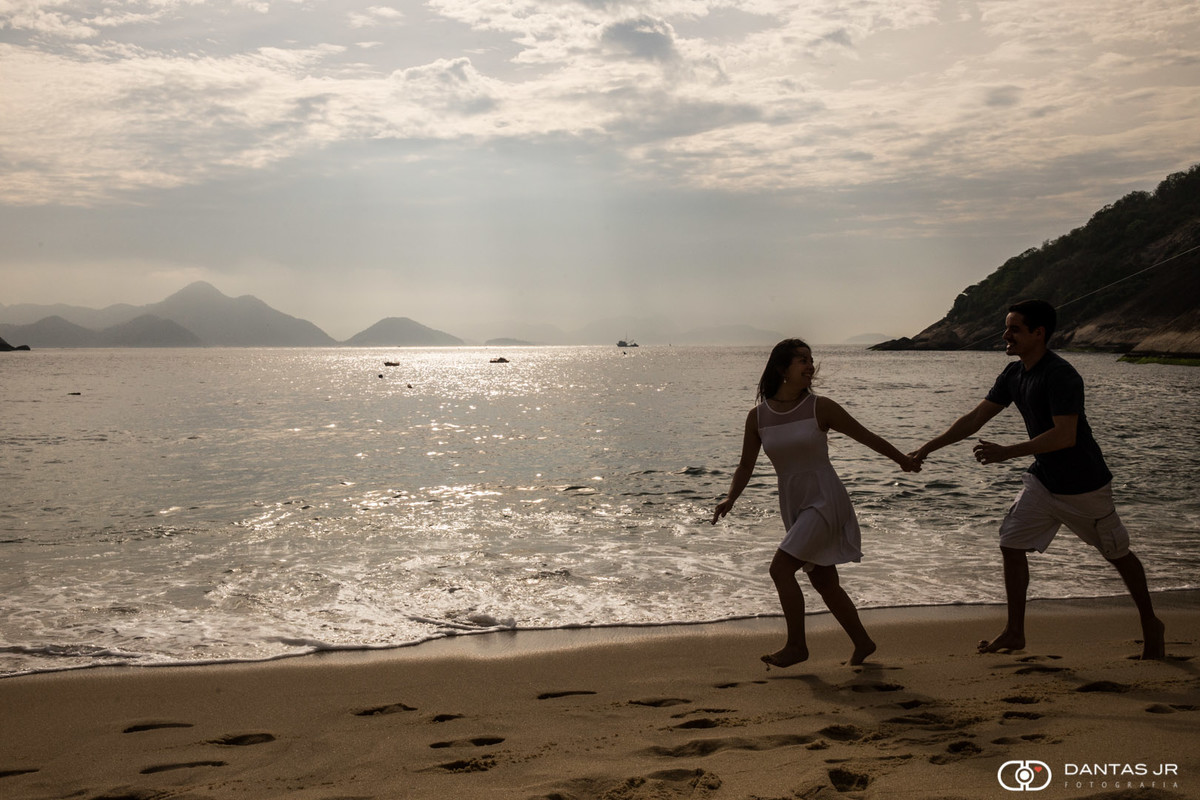casal andando de mãos dadas na praia com linda luz e quase em silhueta apaixonados por Dantas Jr.