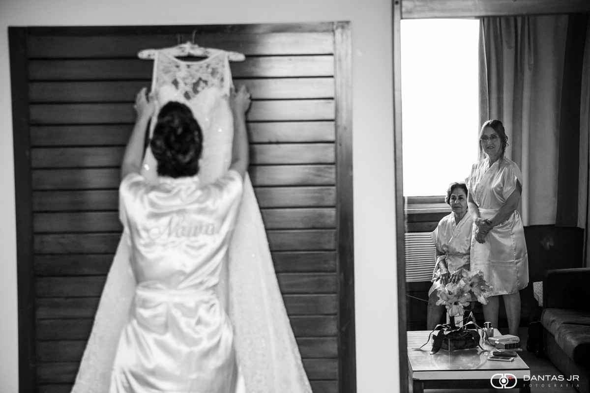 noiva arrumando vestido pendurado no quarto durante making of observada pela mãe e avó através do espelho