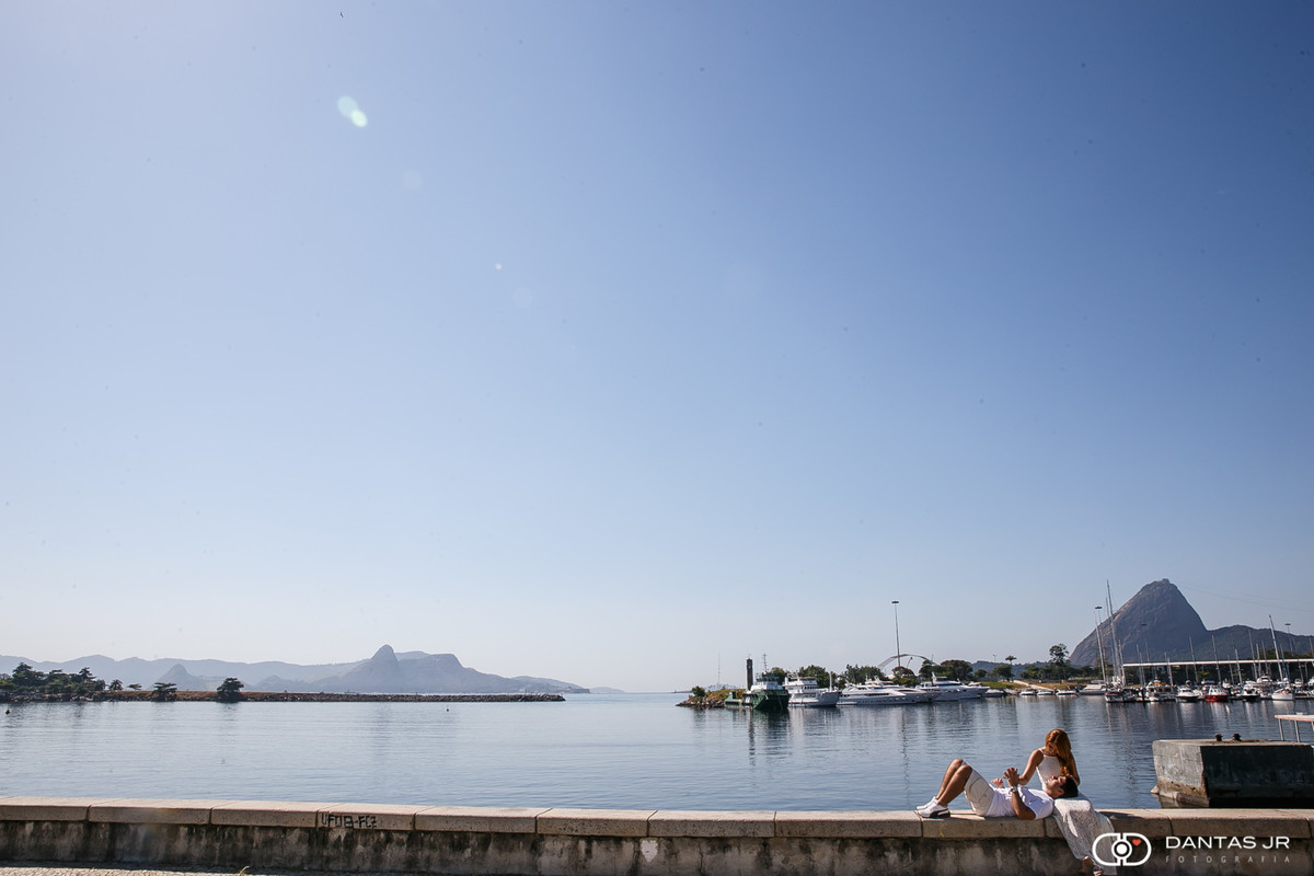 casal sentado em mureta com a urca e pão de açucar ao fundo em dia azul namorando em ensaio pre wedding por Dantas Jr. no Rio de Janeiro