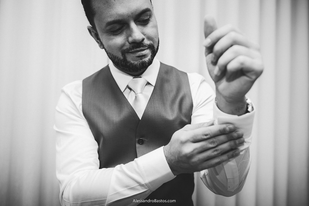 abotoando as mangas da camisa está o noivo do casamento em belo horizonte bh foto e sorrindo com uma cortina no ambiente