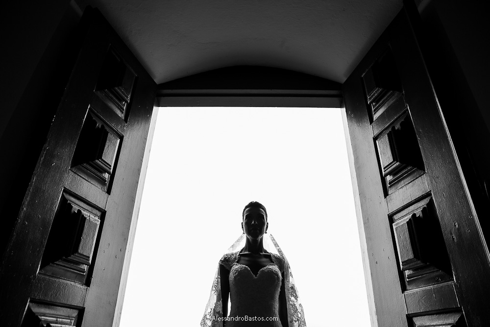 meio corpo da noiva aparece em um contra-luz nas fotografias após o casamento em bh na serra da piedade com as portas da igreja bem aberta