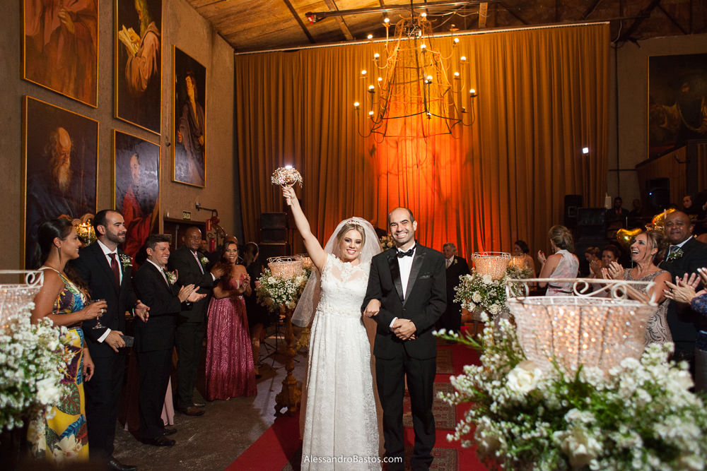 na saída da cerimônia do casamento em bh caravaggio a noiva levanta o seu buquê de broches e o noivos sorriem bem felizes da vida