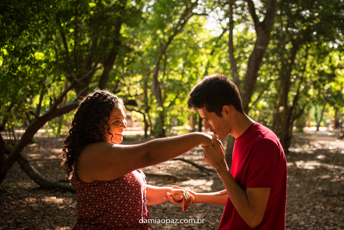 Thiago beijando a mão de Jandsa, no bosque dos namorados, local arborizado e muito ver, com a luz da tarde fazendo um efeito lindo nos dois.
