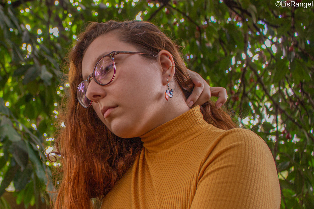 Mulher olhando para a diagonal e para baixo com a mão no cabelo, o fundo contém folhagens.