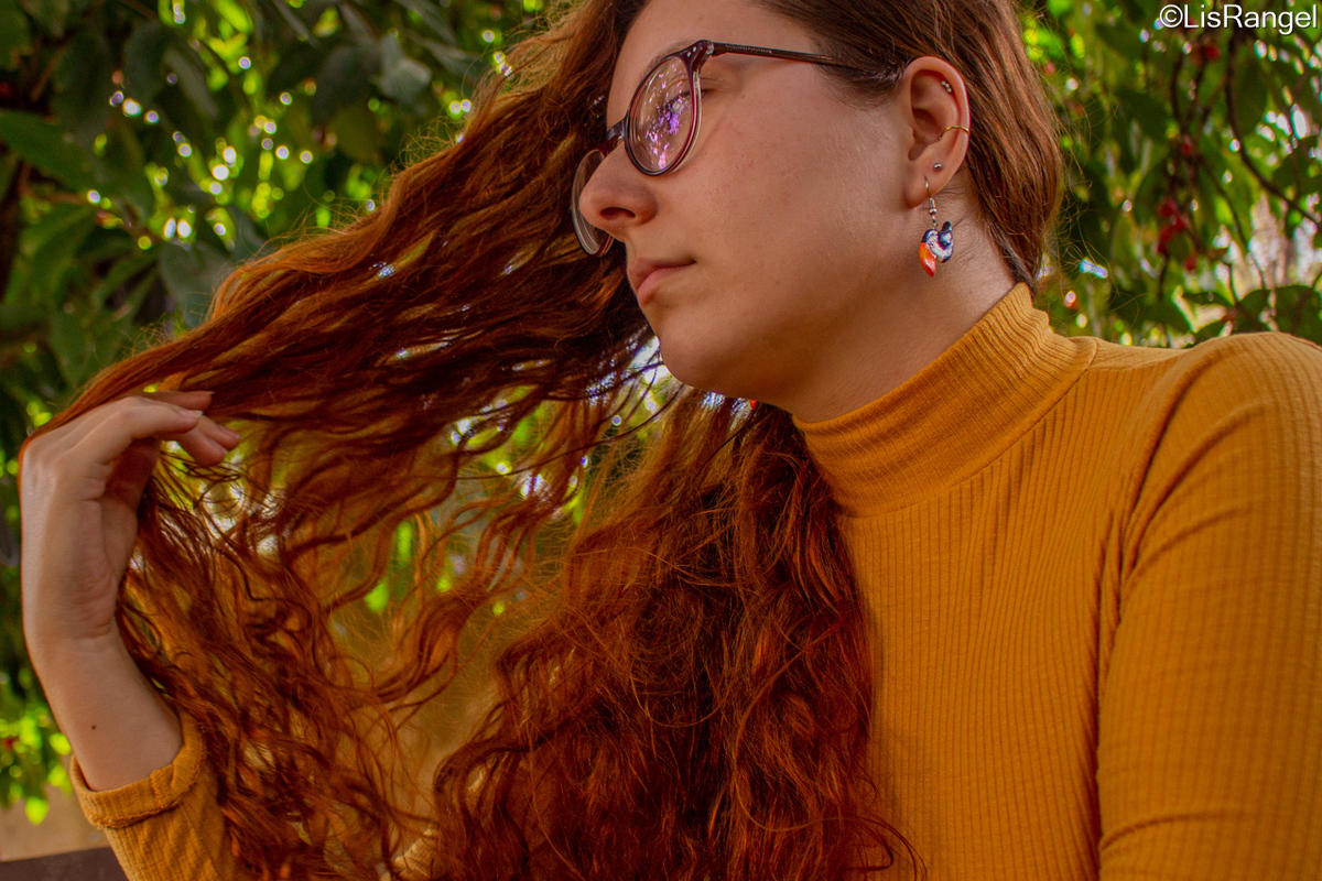Mulher olhando para a diagonal e para baixo com a mão puxando o cabelo, o fundo contém folhagens.