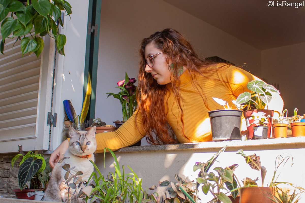 Mulher na janela fazendo carinho em um gato, rodeados de plantas.