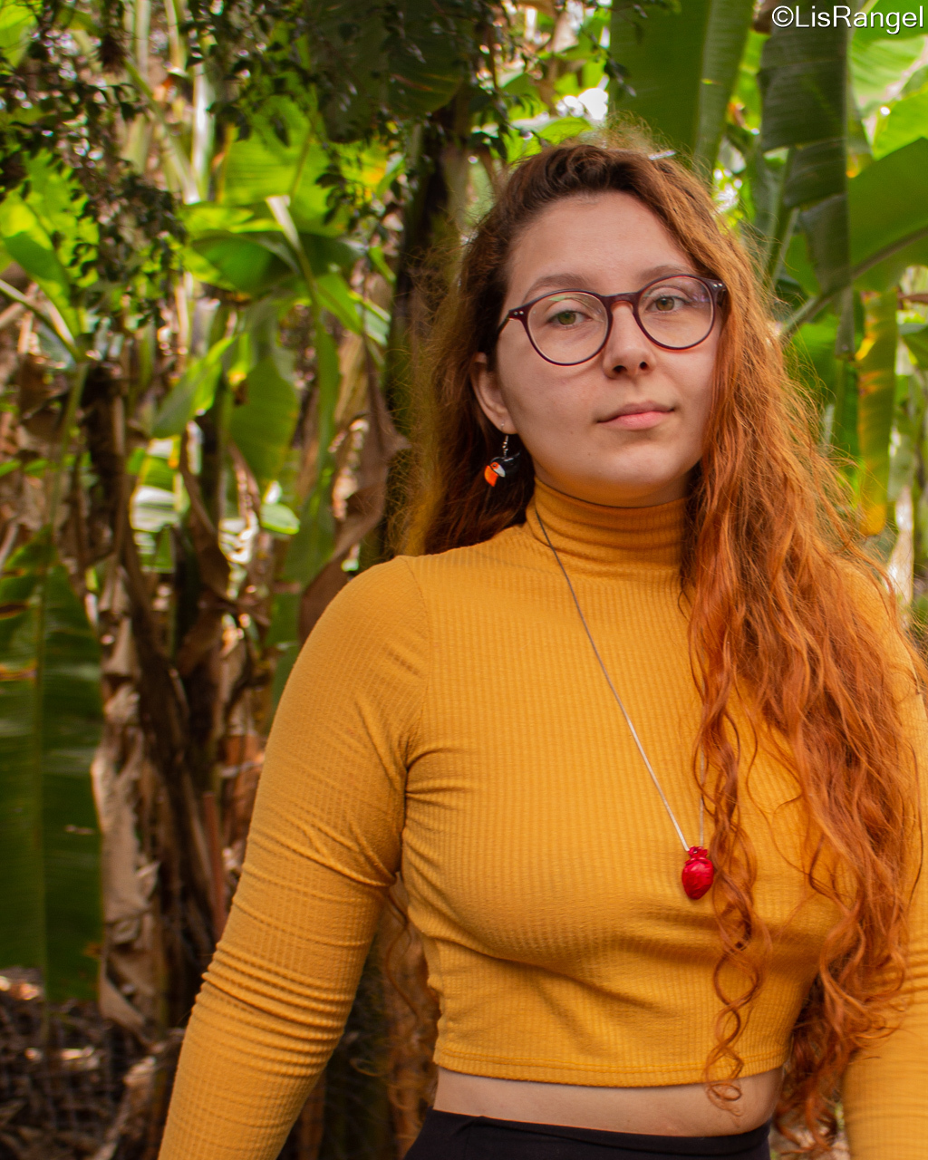 Mulher de frente, olhando para a foto e sorrindo levemente, o fundo contém folhagens.