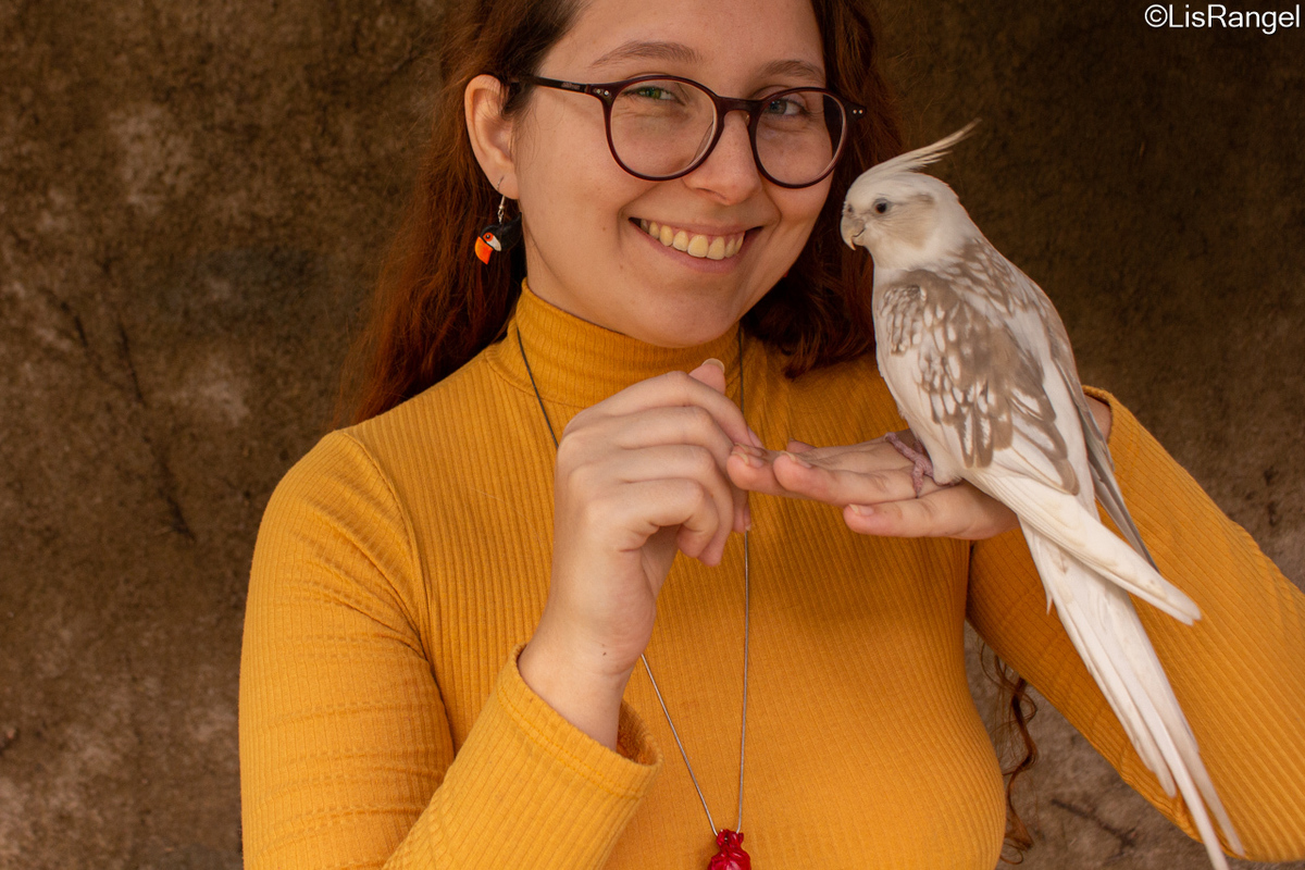 Mulher de frente, olhando para a frente e sorrindo, ela segura uma calopsita, o fundo um muro.