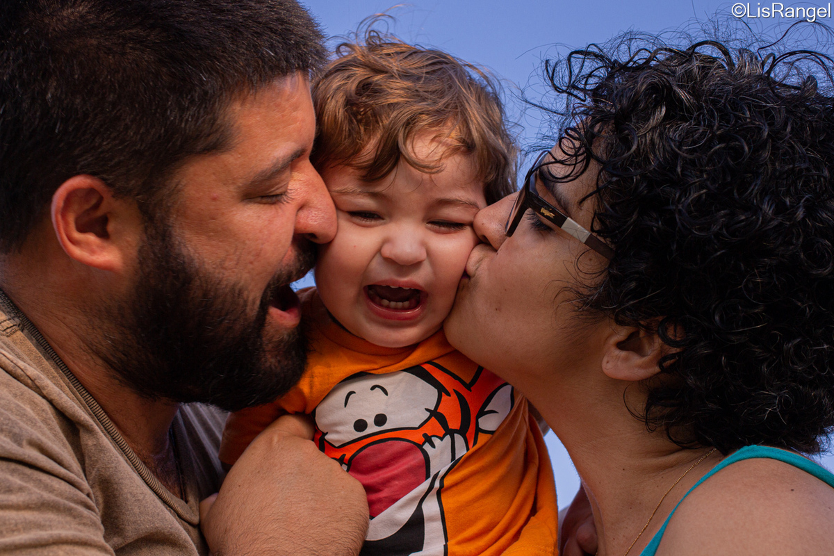 Pai segurando filho que está o rosto amassado, porque a mãe lhe dá um beijo da bochecha.