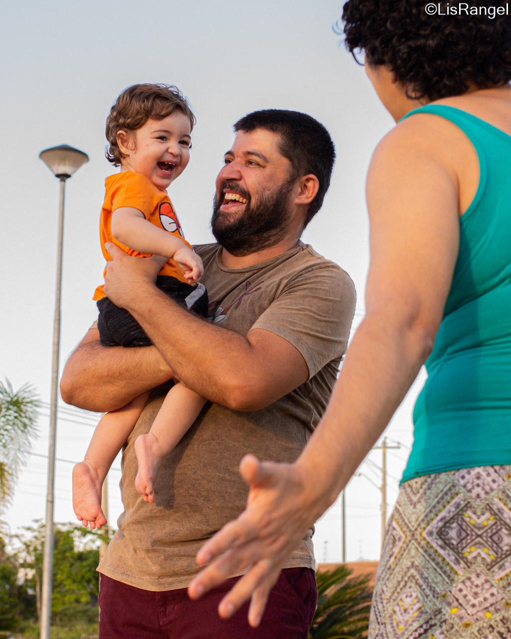 Pai segurando filho que está sorrindo e olha para a mãe que está na frente.