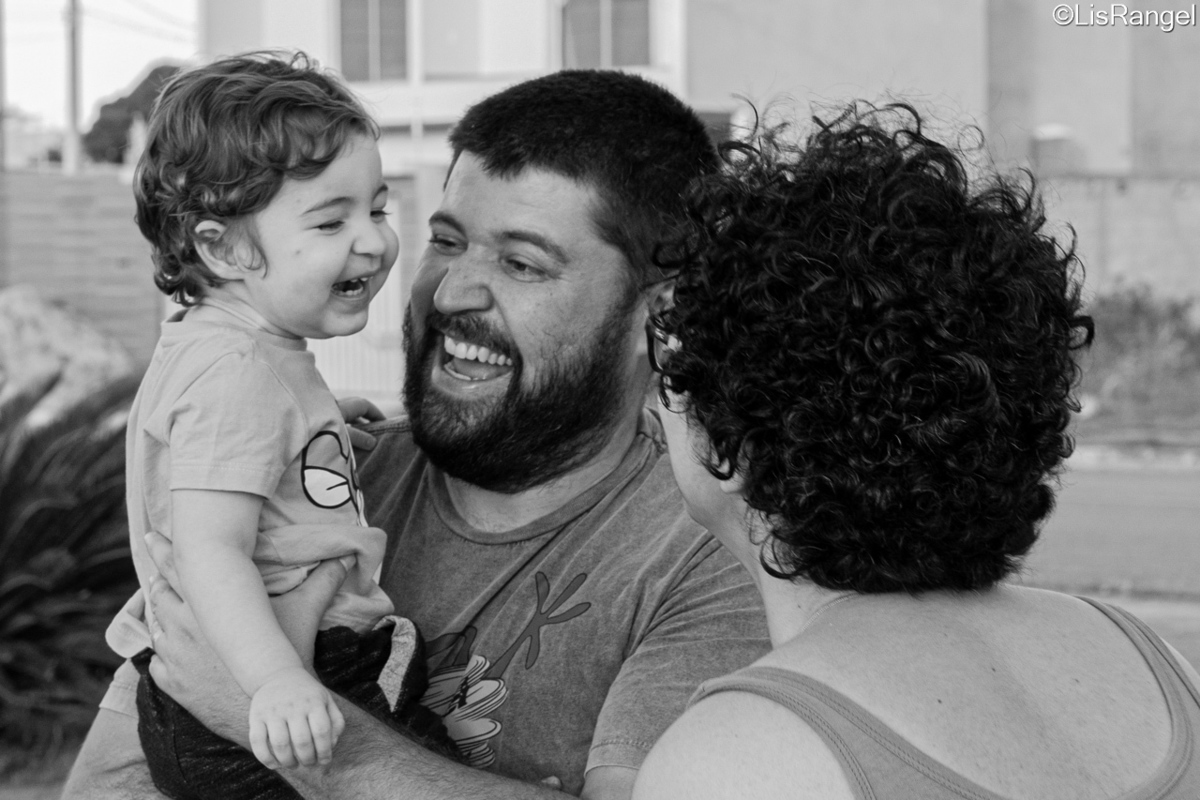 Pai segurando filho que está sorrindo e olha para a mãe que está na frente.