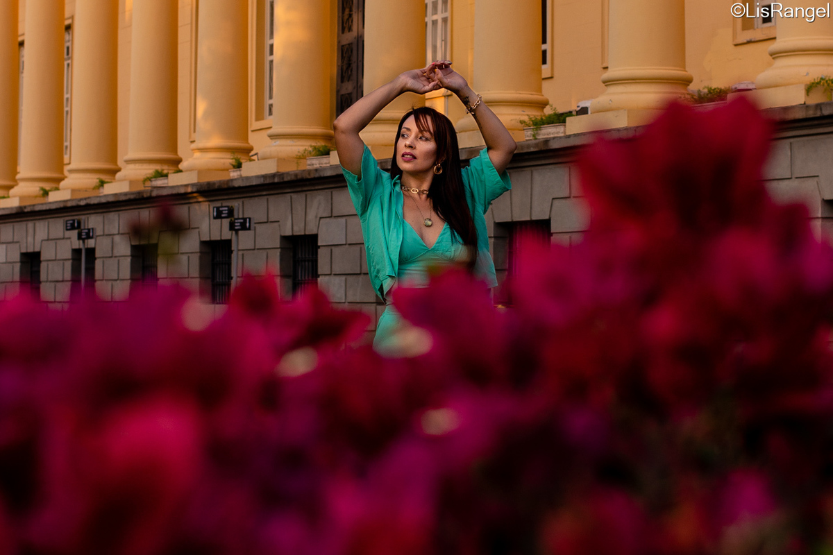 Mulher com as mãos encostadas levemente uma na outra em cima da cabeça, olhando para a esquerda. No primeiro plano em frente da mulher tem flores embaçadas. Veste uma roupa verde clara.