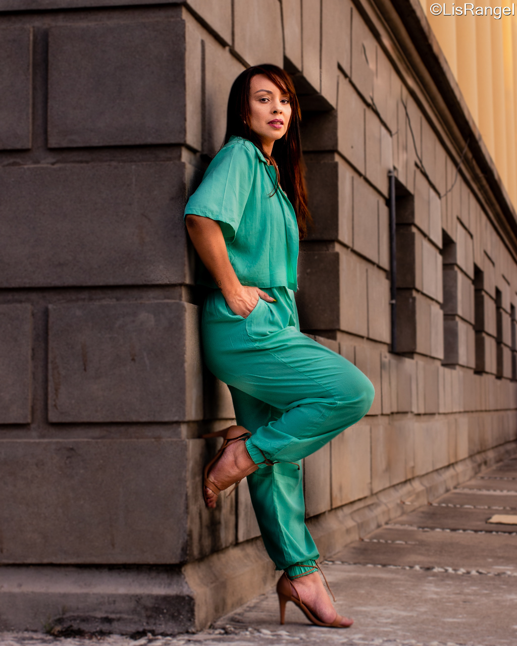 Mulher encostada numa parede, olhando para a câmera. Ela está com um dos pés na parede e as mãos no bolso. Veste uma roupa verde clara.