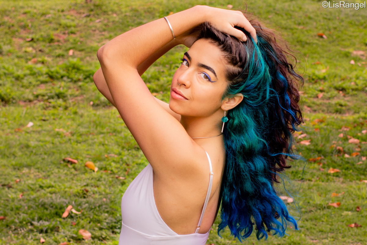 Mulher de lado segurando o cabelo, revelando a parte de baixo do cabelo que está pintada de azul, ela dá um sorriso leve de canto de boca e olha para a câmera.
