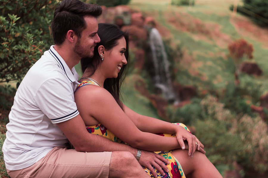 ENSAIO FOTOGRÁFICO PRÉ-WEDDING PRE WEDDING PREWEDDING DE CASAL PARQUE DO JAPAO MARINGA PARANA NA FOTO O CASAL SENTADO NO CHAO OBSERVA O HORIZONTE E NO FUNDO UMA CACHOEIRA