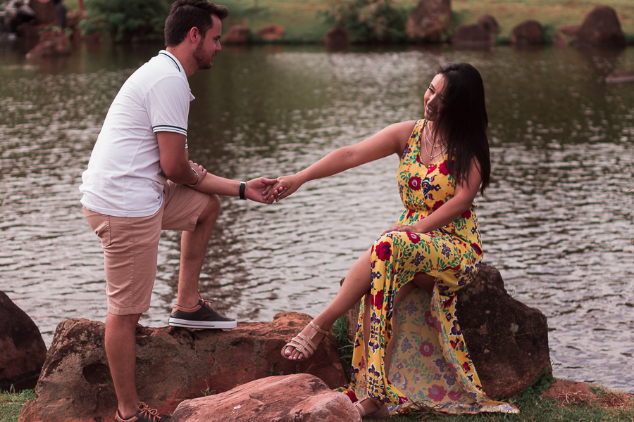ENSAIO FOTOGRÁFICO PRÉ-WEDDING PRE WEDDING PREWEDDING DE CASAL PARQUE DO JAPAO MARINGA PARANA NA FOTO A NOIVA APARECE SENTADA A BEIRA DO LAGO ENQUANTO SEU NOIVO SEGURA A SUA MAO