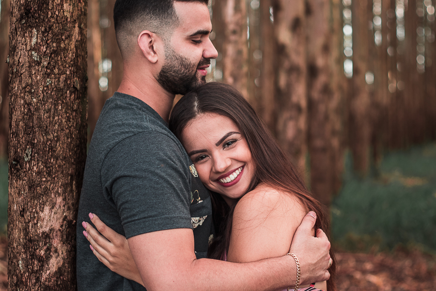 ENSAIO PRE WEDDING MANDAGUACU PARANA PRE CASAMENTO MARINGA CASAL NO EUCALIPTO ELE ESTA ENCOSTADO NA ARVORE ELA SORRINDO OLHANDO PARA A CAMERA 