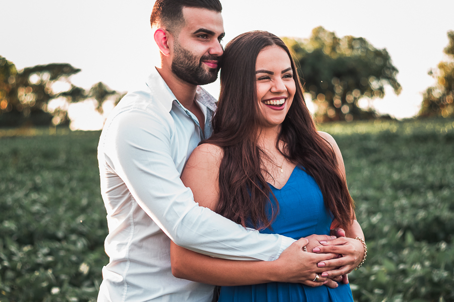 ENSAIO PRE WEDDING MANDAGUACU PARANA PRE CASAMENTO MARINGA O CASAL ESTA EM UMA PLANTAÇAO ELE ESTA SERIO OLHANDO PARA O HORIZONTE ABRAÇANDO A CINTURA DELA E ELA ESTA COM O SORRISO ESPONTANEO E COM AS MAOS ENTRELACADAS COM AS DELE