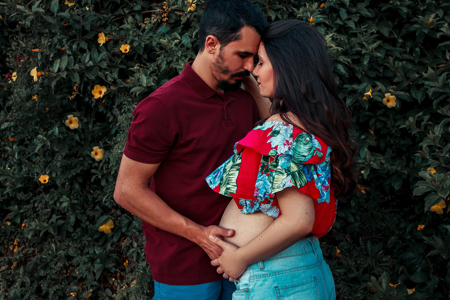 ENSAIO FOTOGRAFICO DE GESTANTE GESTACAO REALIZADO EM MARINGA NO PARANA NA IMAGEM O CASAL ESTA COM AS MAOS NA BARRIGA ENQUANTO ESTAO COM AS TESTAS COLADAS E OLHOS FECHADOS EM SINAL DE CARINHO E REFLEXAO VARIAS FLORES AMARELAS DE FUNDO