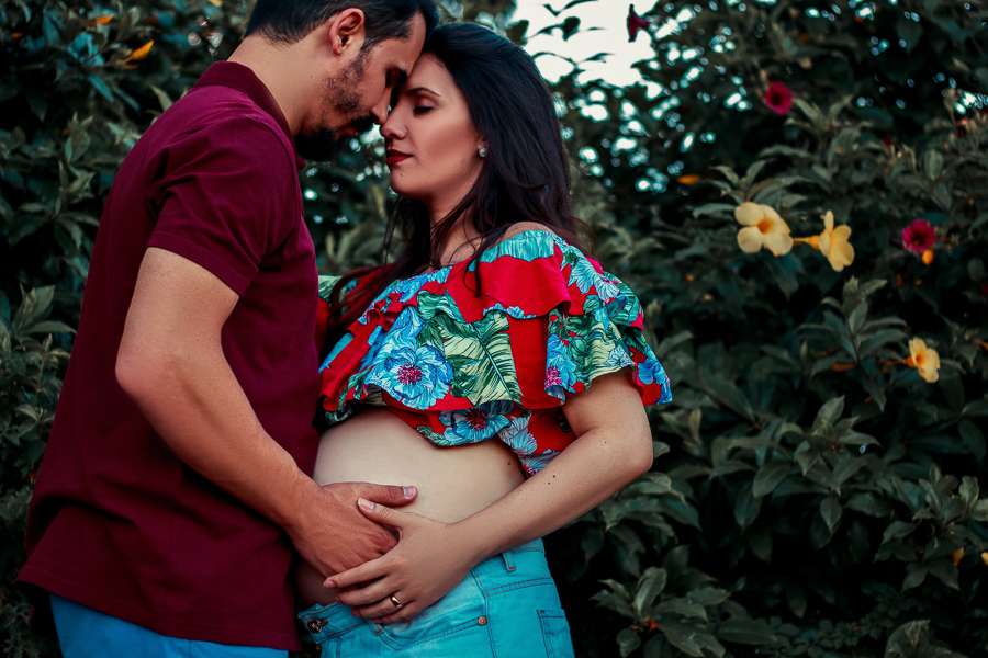 ENSAIO FOTOGRAFICO DE GESTANTE GESTACAO REALIZADO EM MARINGA NO PARANA NA IMAGEM O CASAL ESTA COM AS MAOS NA BARRIGA ENQUANTO ESTAO COM AS TESTAS COLADAS E OLHOS FECHADOS EM SINAL DE CARINHO E REFLEXAO VARIAS FLORES AMARELAS E VERMELHAS NO FUNDO