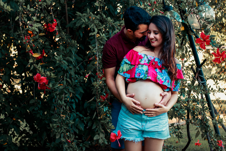 ENSAIO FOTOGRAFICO DE GESTANTE GESTACAO REALIZADO EM MARINGA NO PARANA NA IMAGEM OS PAPAIS ESTAO ABRAÇADOS E COM AS MAOS NA BARRIGA ENQUANTO O PAPAI DA UM CHEIRO NA MAMAE ELA SORRI COM VARIAS FLORES VERMELHAS DE FUNDO