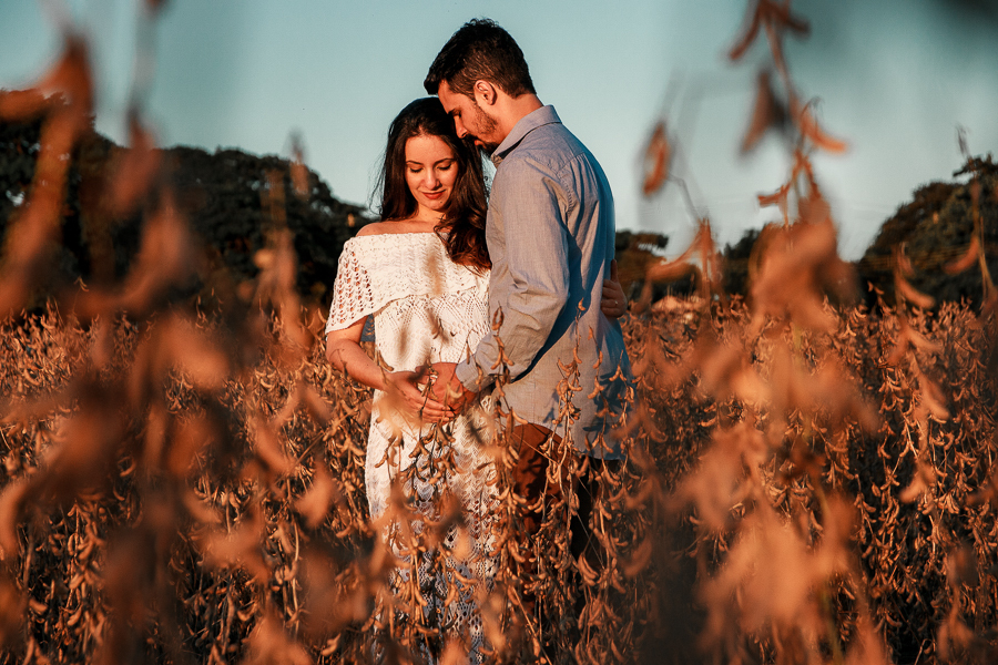 ENSAIO FOTOGRAFICO DE GESTANTE GESTACAO REALIZADO EM MARINGA NO PARANA NA IMAGEM A MAMAE APARECE COM UM VESTIDO BRANCO ENQUANTO O PAI SEGURA A BARRIGA DELA COM CARINHO FOTO EM MEIO A SOJAS NO PRIMEIRO PLANO