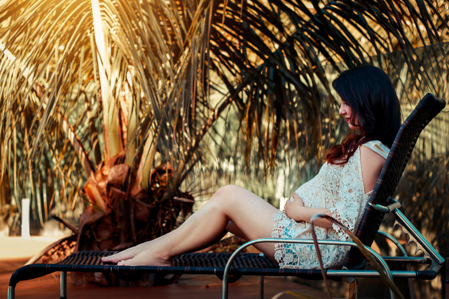ENSAIO FOTOGRAFICO DE GESTANTE GESTACAO REALIZADO EM MARINGA NO PARANA NA IMAGEM A FUTURA MAMÃE SEGURA SUA BARRIGA ENQUANTO ESTA SENTADA EM UMA CONFORTÁVEL CADEIRA COM FUNDO UMA PALMEIRA