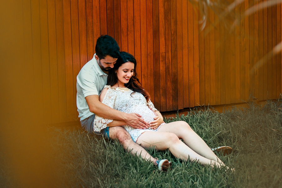 ENSAIO FOTOGRAFICO DE GESTANTE GESTACAO REALIZADO EM MARINGA NO PARANA NA IMAGEM OS FUTUROS PAPAIS ESTAO SENTADOS E ENCOSTADOS EM UM DECK DE MADEIRA ENQUANTO A MAMAE SORRI O PAPAI SEGURA A BARRIGA