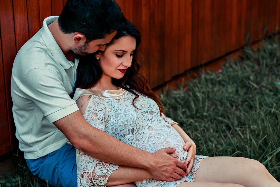 ENSAIO FOTOGRAFICO DE GESTANTE GESTACAO REALIZADO EM MARINGA NO PARANA NA IMAGEM OS FUTUROS PAPAIS ESTAO SENTADOS E ENCOSTADOS EM UM DECK AMADEIRADO ENQUANTO O PAPAI SEGURA A BARRIGA DA MAMAE ELA OLHA PARA BAIXO NUM SINAL DE CARINHO