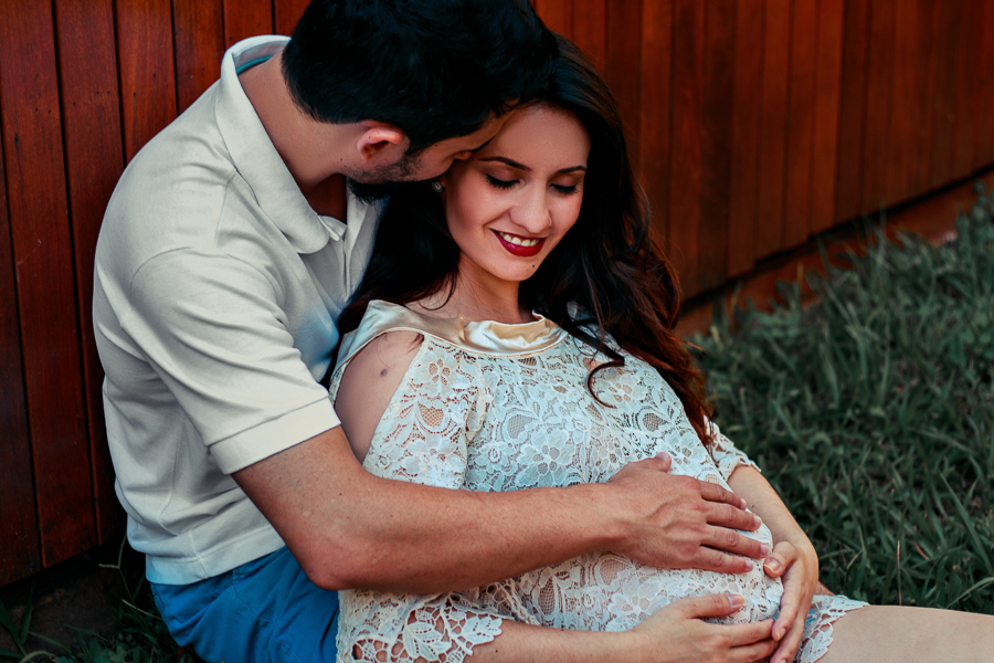 ENSAIO FOTOGRAFICO DE GESTANTE GESTACAO REALIZADO EM MARINGA NO PARANA NA IMAGEM OS FUTUROS PAPAIS ESTAO SENTADOS E ENCOSTADOS EM UM DECK DE MADEIRA AVERMELHADA O PAPAI FAZ CARINHO NA BARRIGA ENQUANTO CHEIRA E DA BEIJOS NO CABELO DA MAMAE QUE SORRI